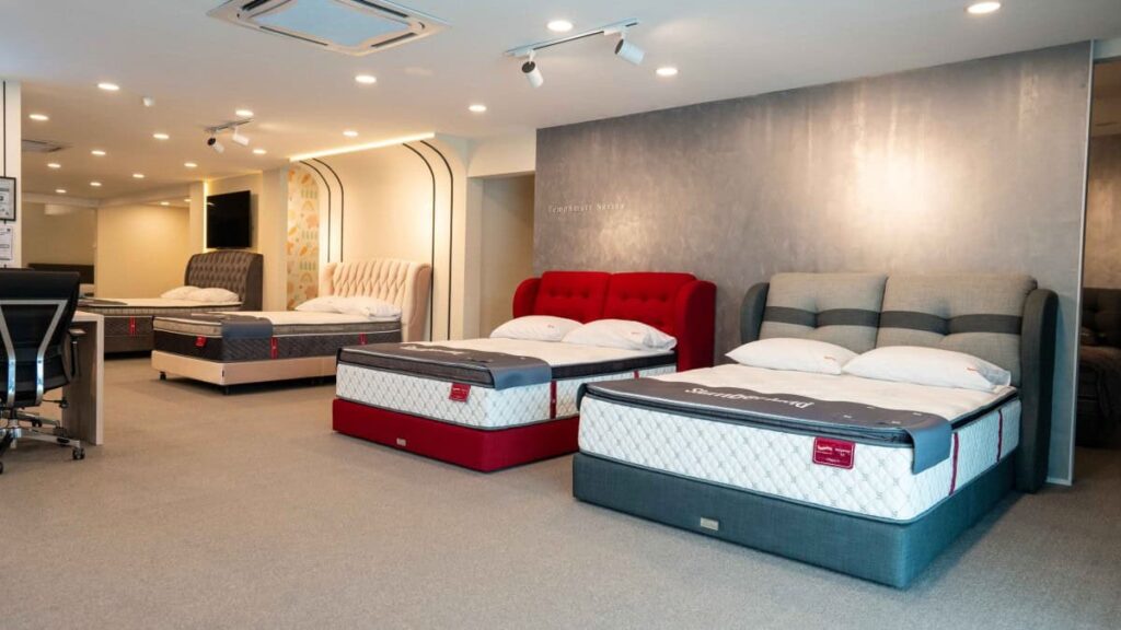 Interior of a mattress store displaying several beds with different upholstered headboards and mattress designs. The well-lit showroom features gray carpeting, modern decor, and a comfortable layout that showcases Slumberland Malaysia mattress options for customers.